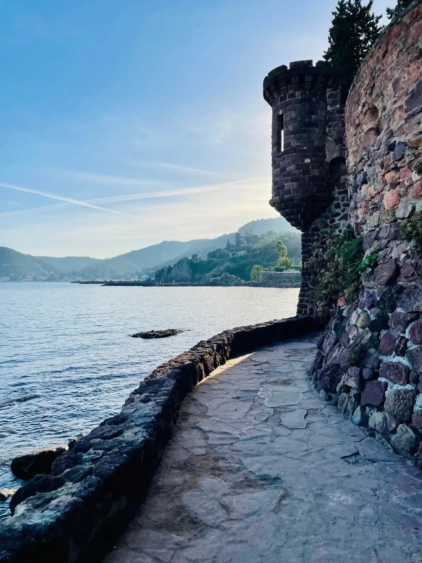 Mur de pierre ancien longeant un chemin pavé au bord de l'eau avec une tour ronde et des montagnes en arrière-plan