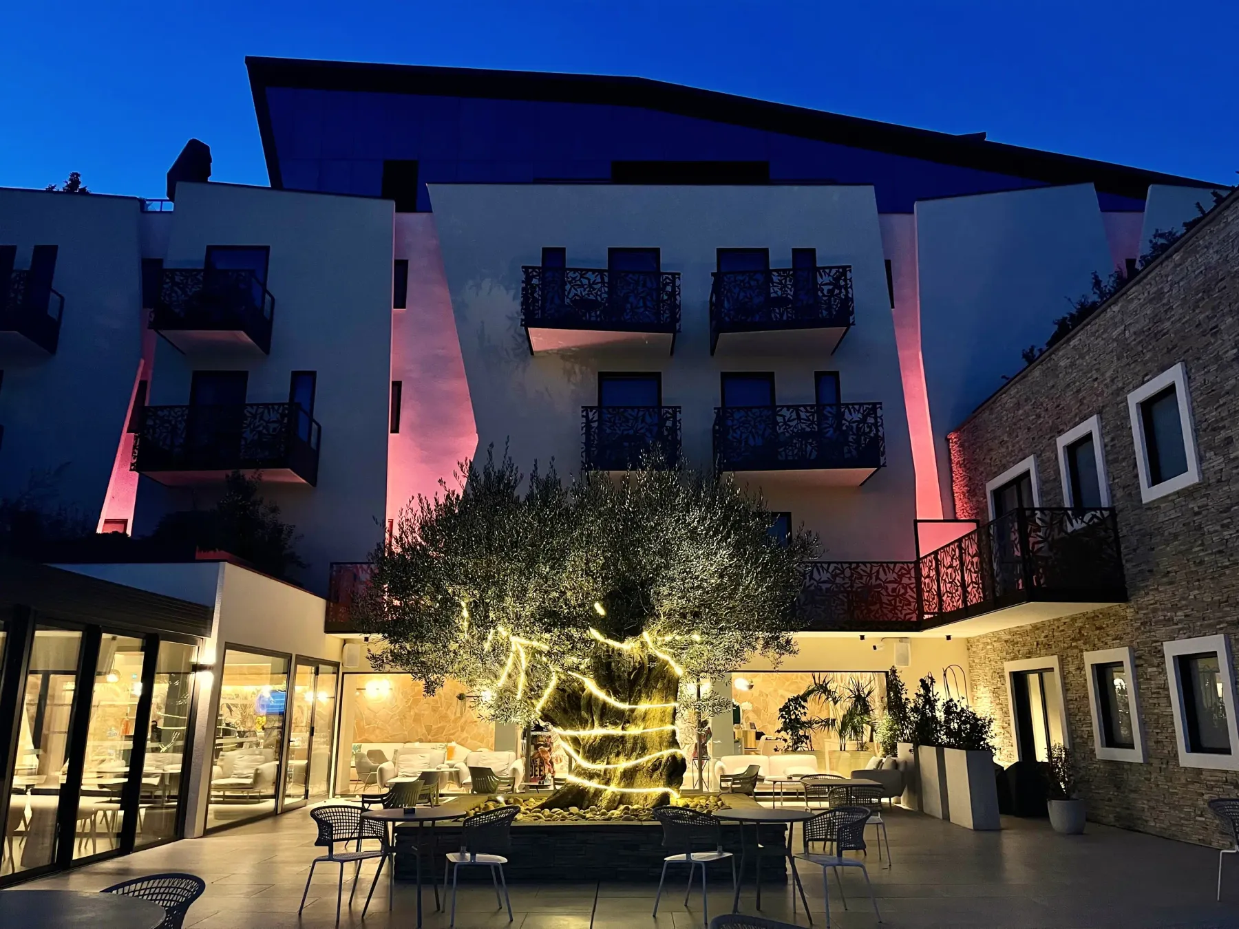 Cour intérieure d'un bâtiment moderne avec des balcons, éclairée la nuit, contenant des tables, des chaises et un arbre illuminé au centre