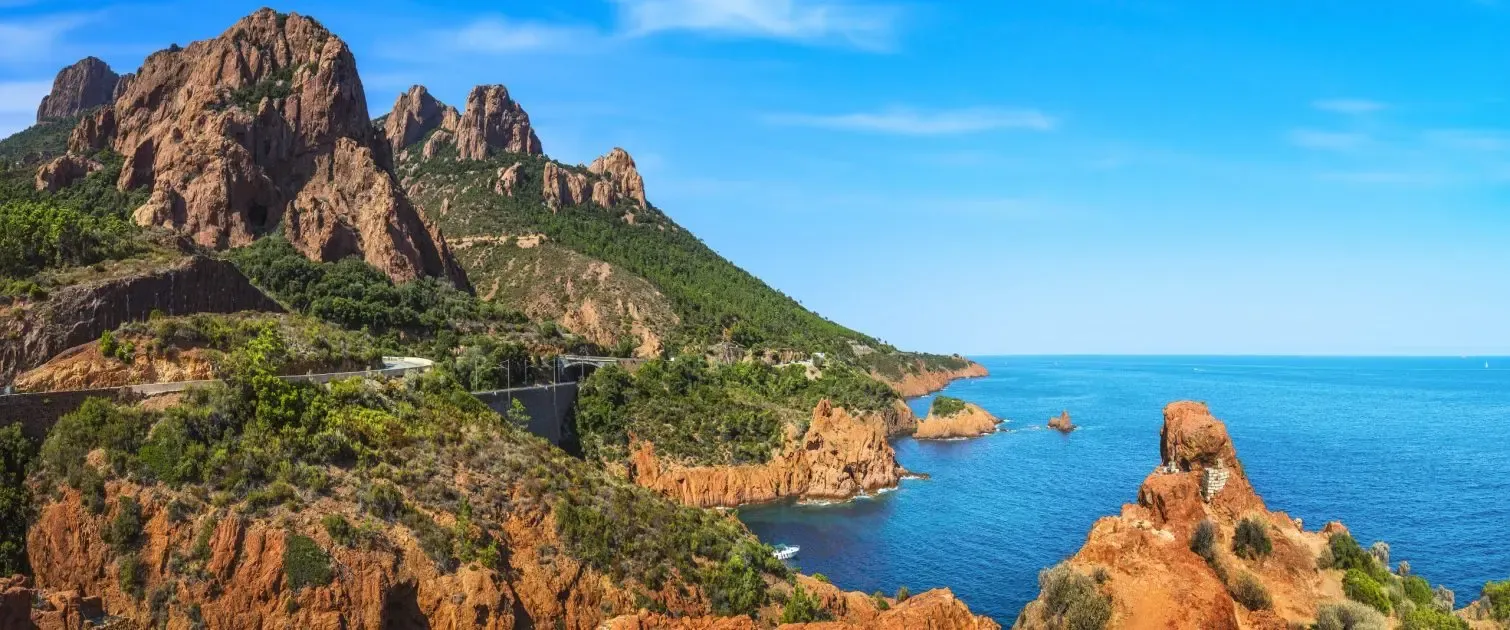 A rocky coastline with steep mountain formations and a winding road along the blue sea under a clear sky