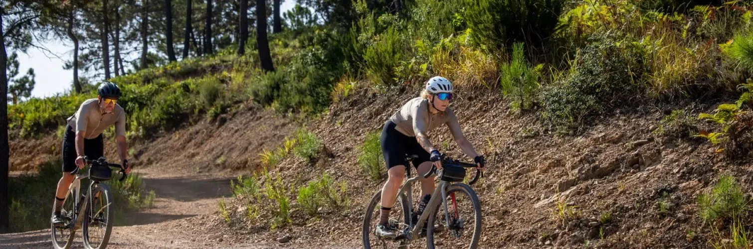 Two cyclists are wearing helmets and sunglasses; they are riding on a dirt path in the forest.
