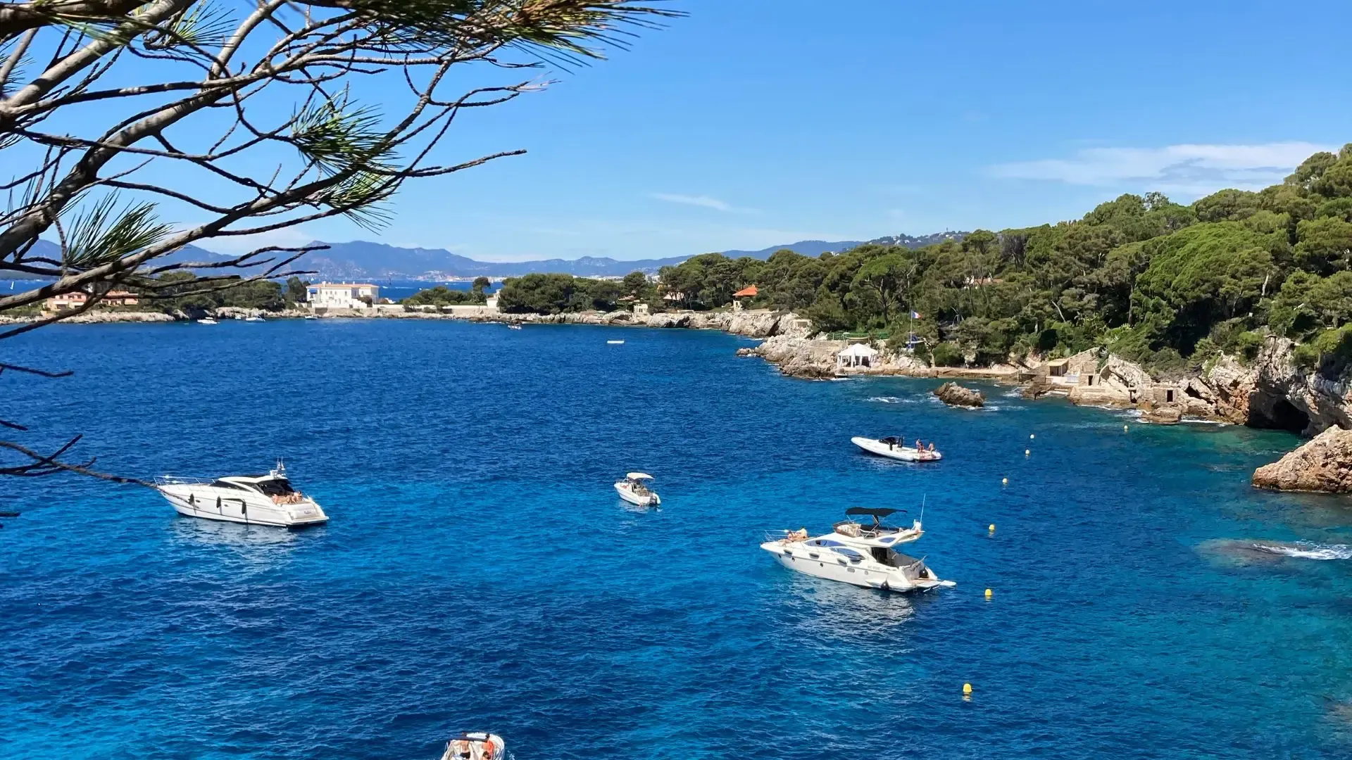 Several boats sail in a turquoise bay bordered by vegetation and rocks, with mountains in the background.
