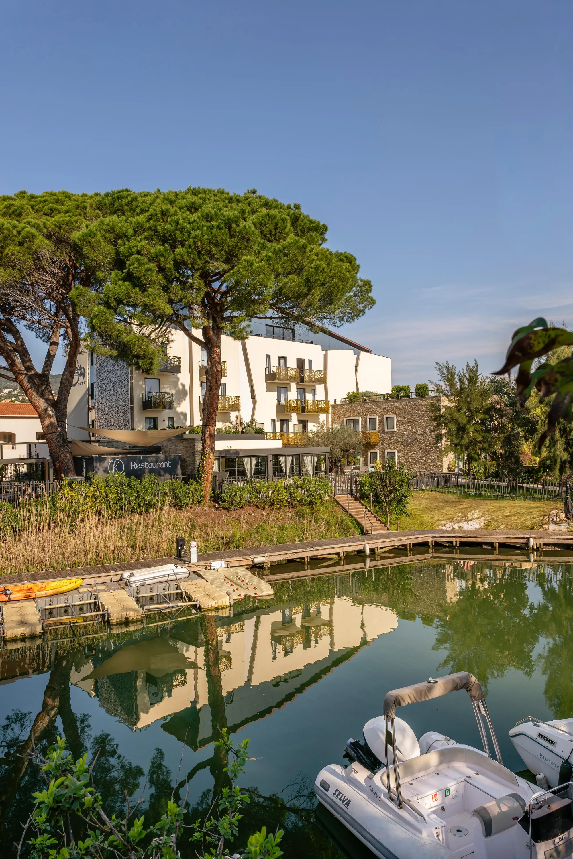 A canal with moored boats, reflecting a white building and trees, surrounded by vegetation and wooden structures.