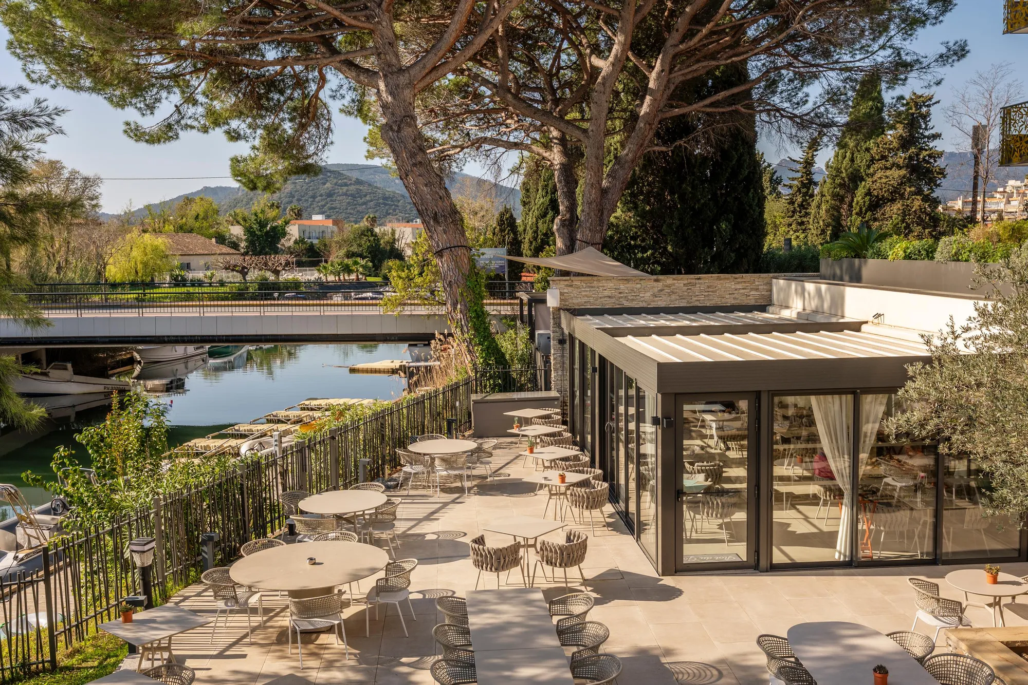 Café terrace with outdoor tables and chairs, lined with trees and a body of water, located near a modern building with large glass windows, with mountains in the background.