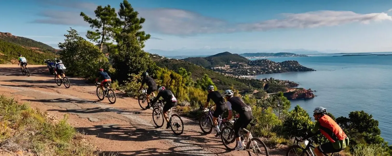 A group of cyclists ride on a dirt path by the sea with a view of a coastal town and hills.