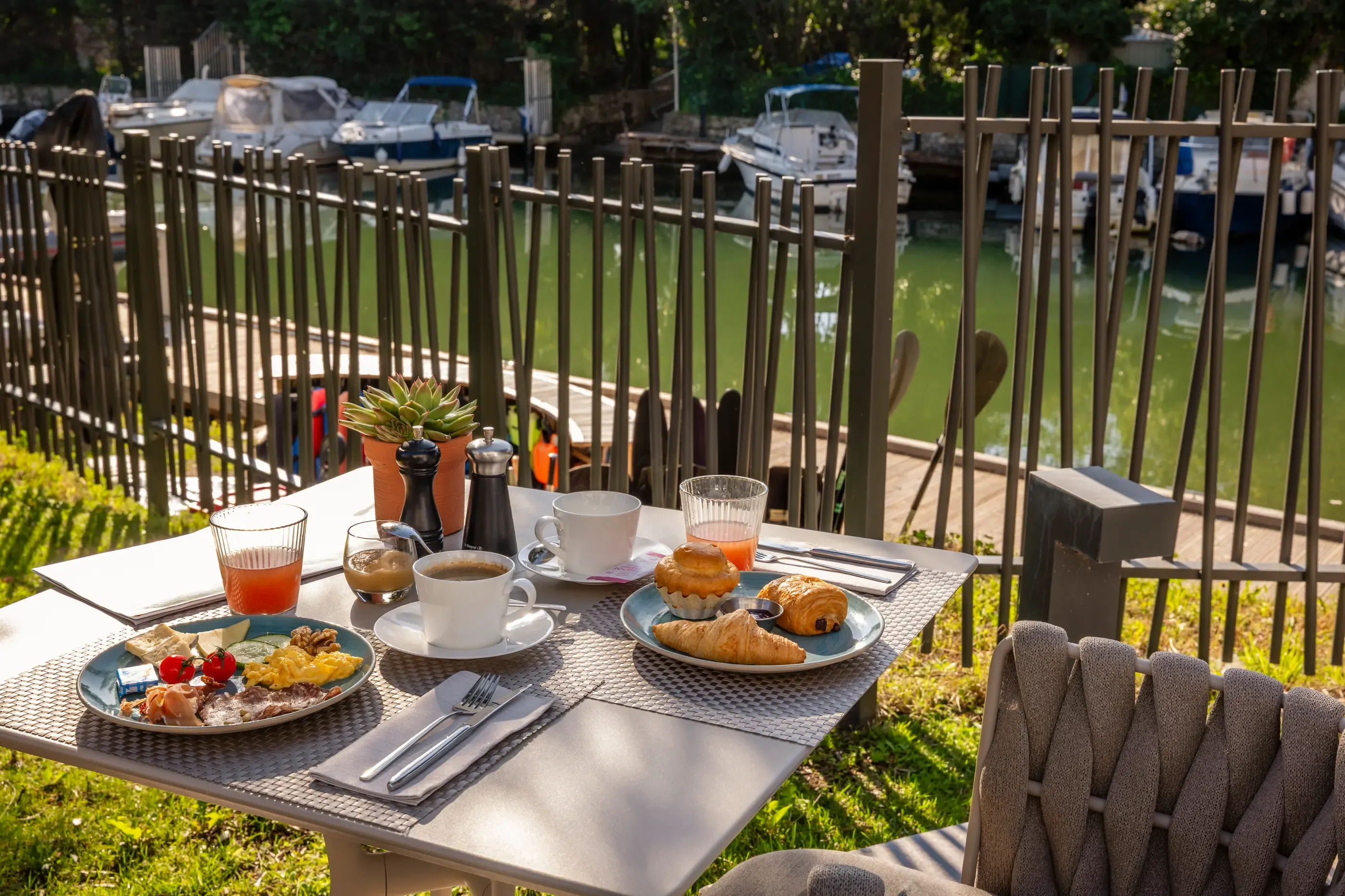 Outdoor table with breakfast consisting of croissants, bread, jam, fruit, and drinks, view of a canal with boats