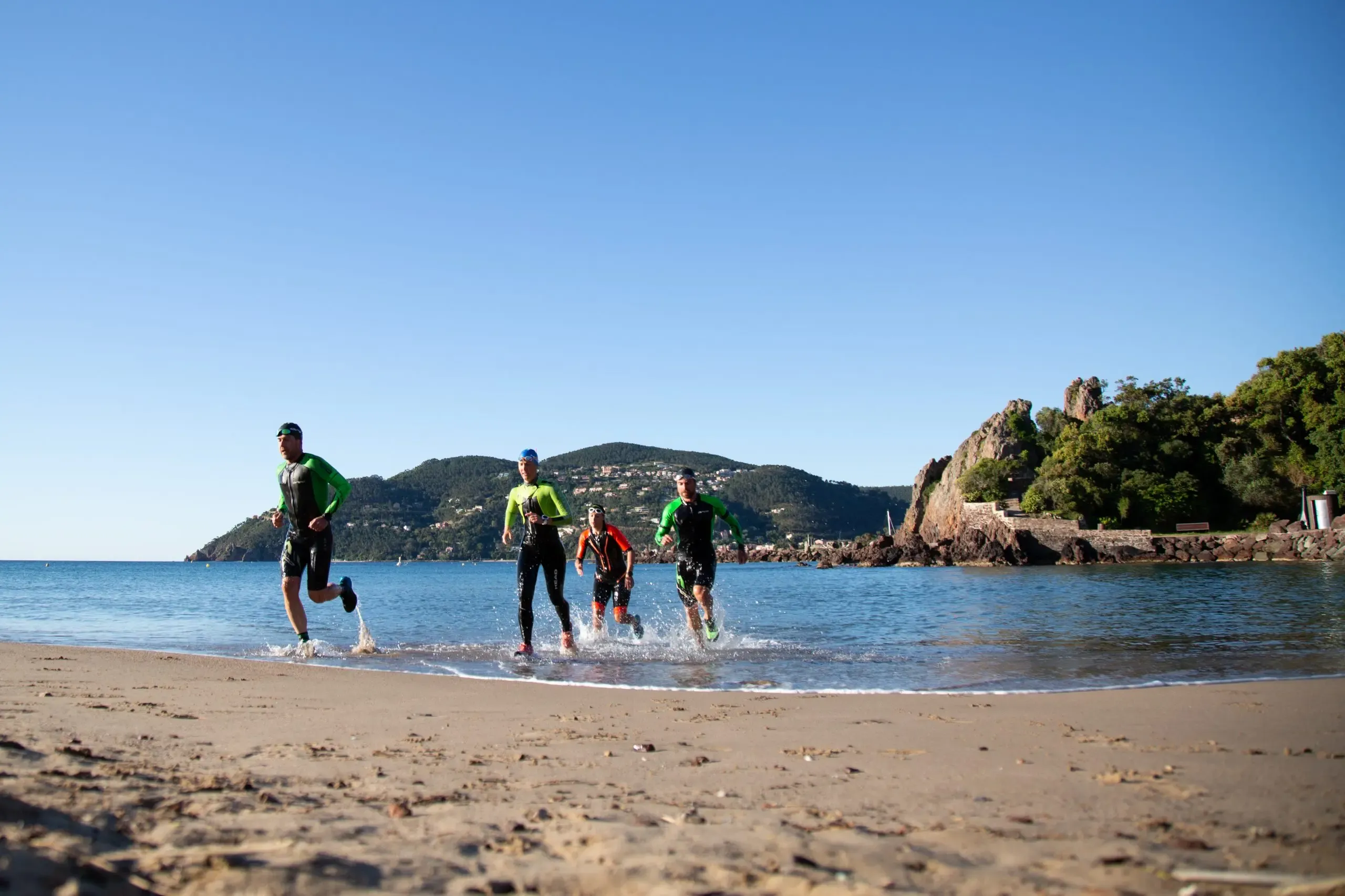 Four people in sportswear are running on a beach with mountains in the background.