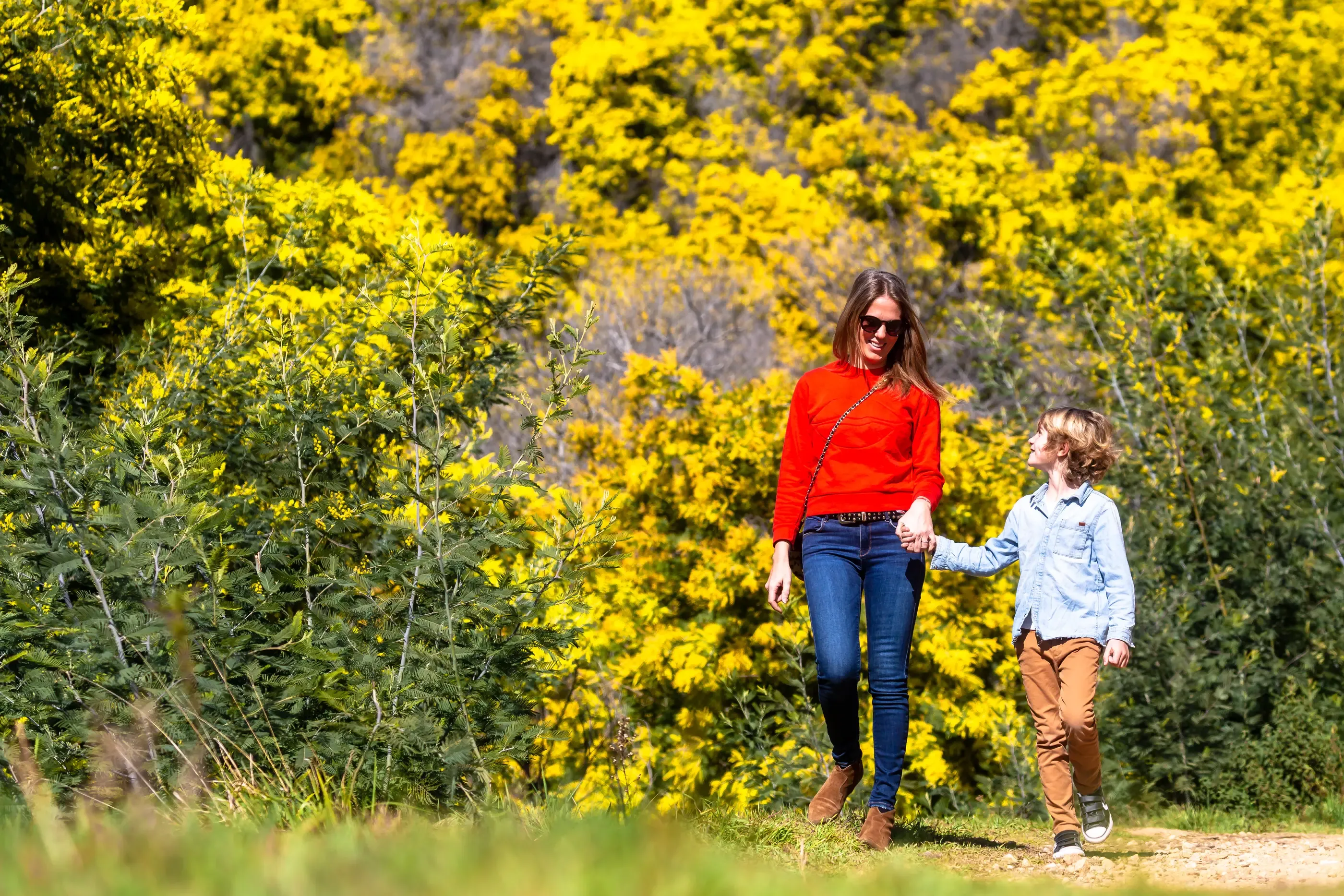 Une femme en rouge et un enfant en bleu marchent main dans la main sur un chemin entouré de végétation jaune