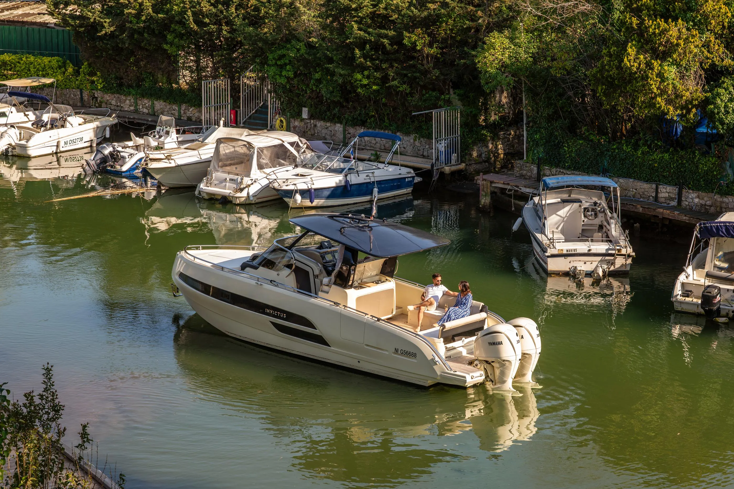 Un bateau de plaisance blanc avec deux personnes à bord est amarré dans un petit port entouré d'autres embarcations et de végétation