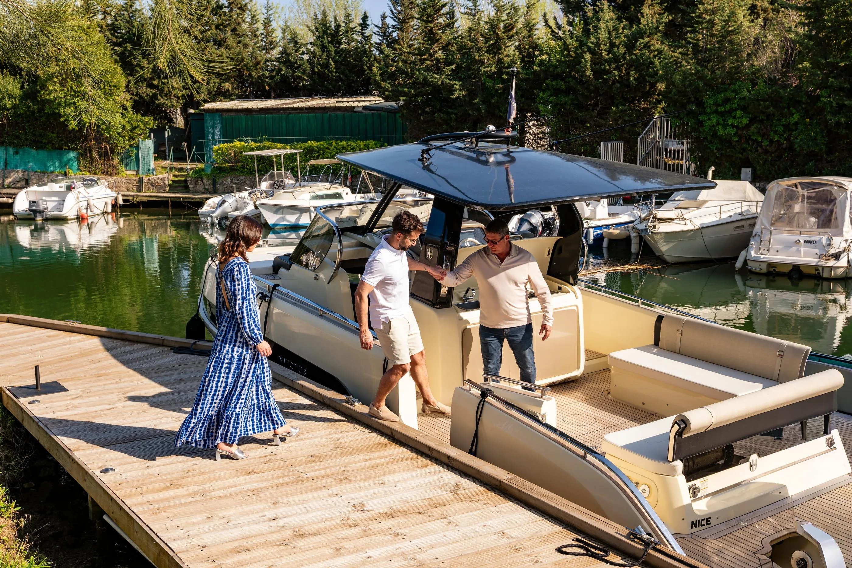 Three people board a boat moored in a port surrounded by other vessels and vegetation.