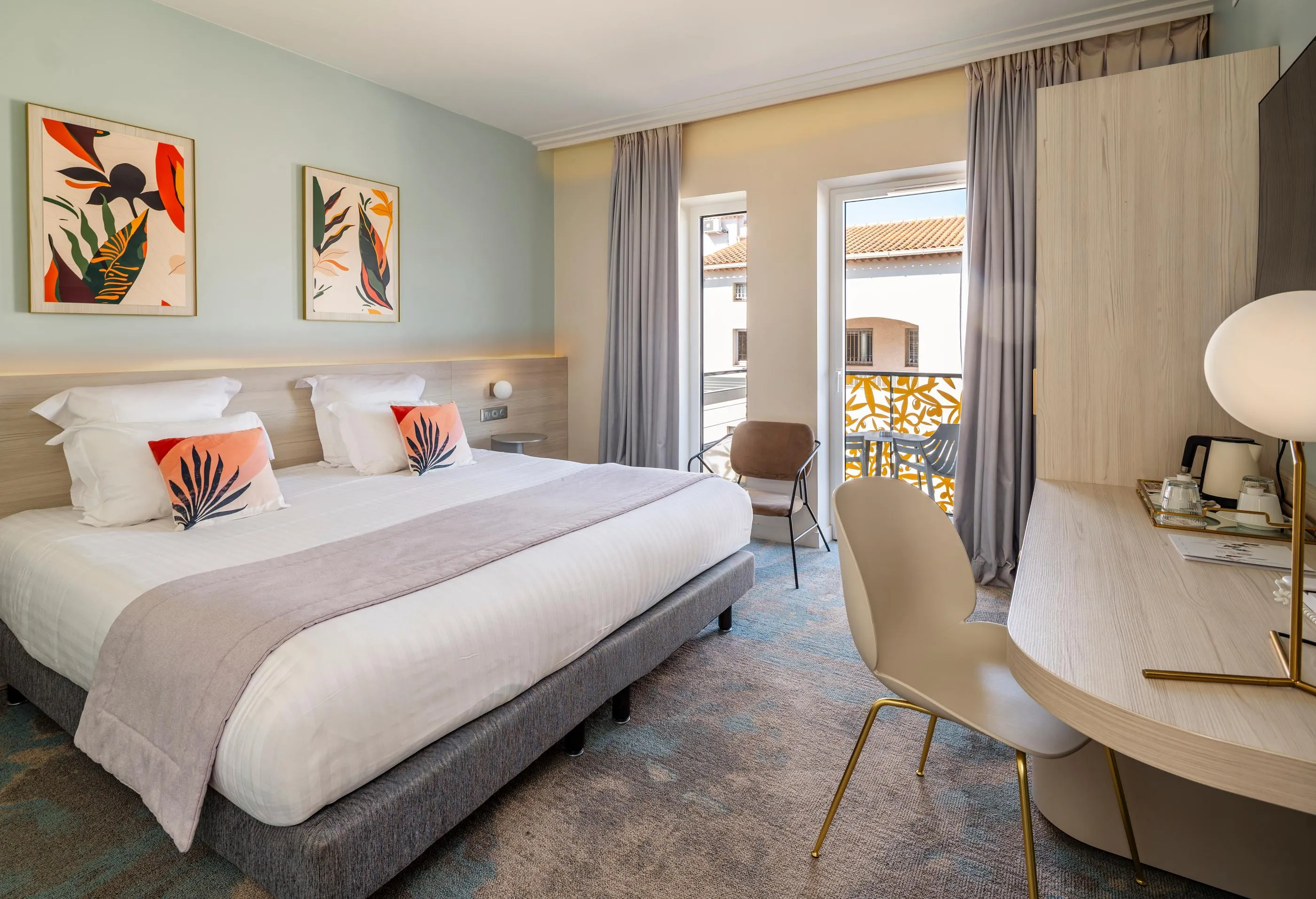 Hotel room with a double bed, a bedside table, a chair, and a French door leading to a balcony with a view of rooftops.