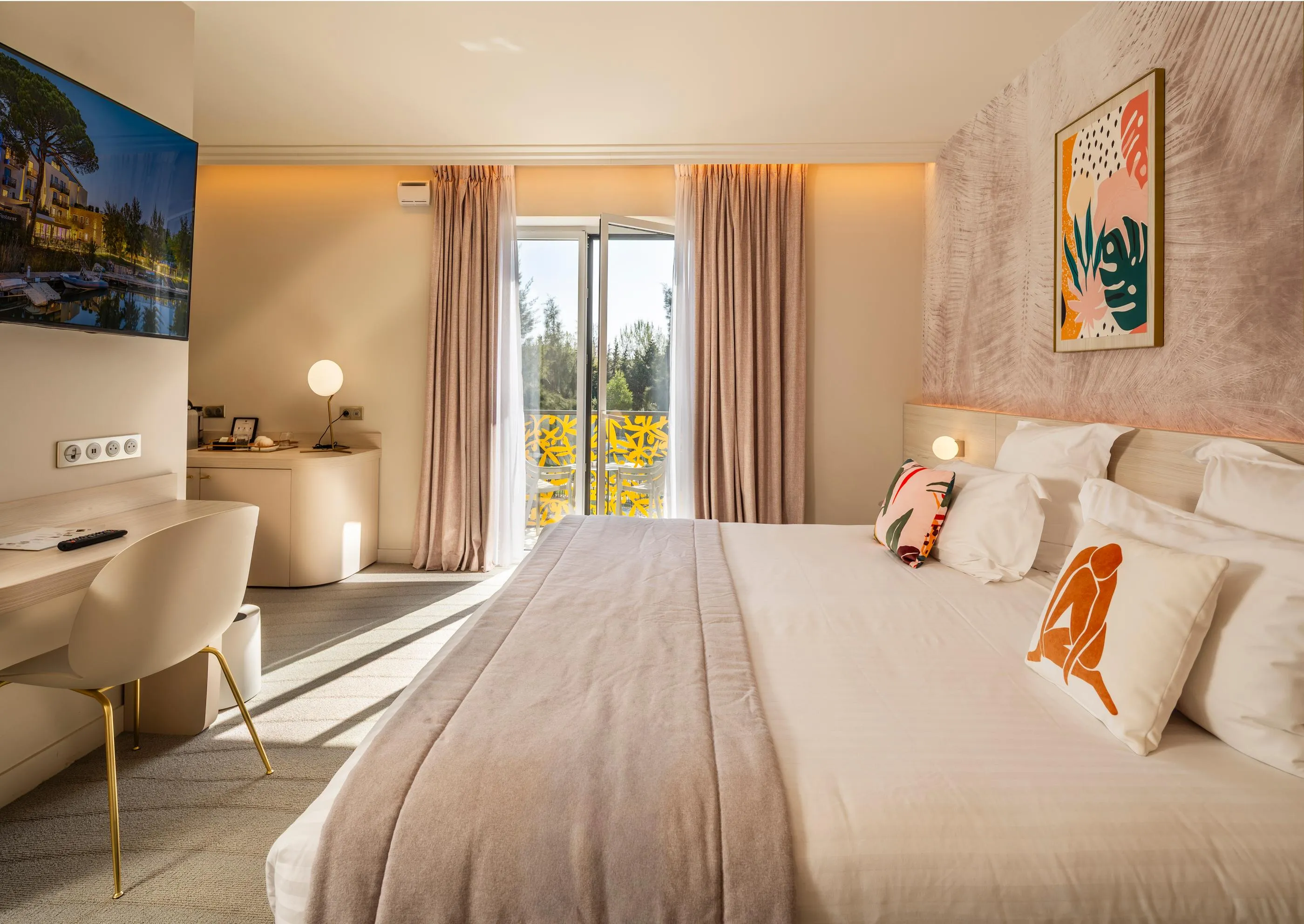 Hotel room with a large bed, a work desk, artwork on the walls, and a sliding door leading to a balcony.