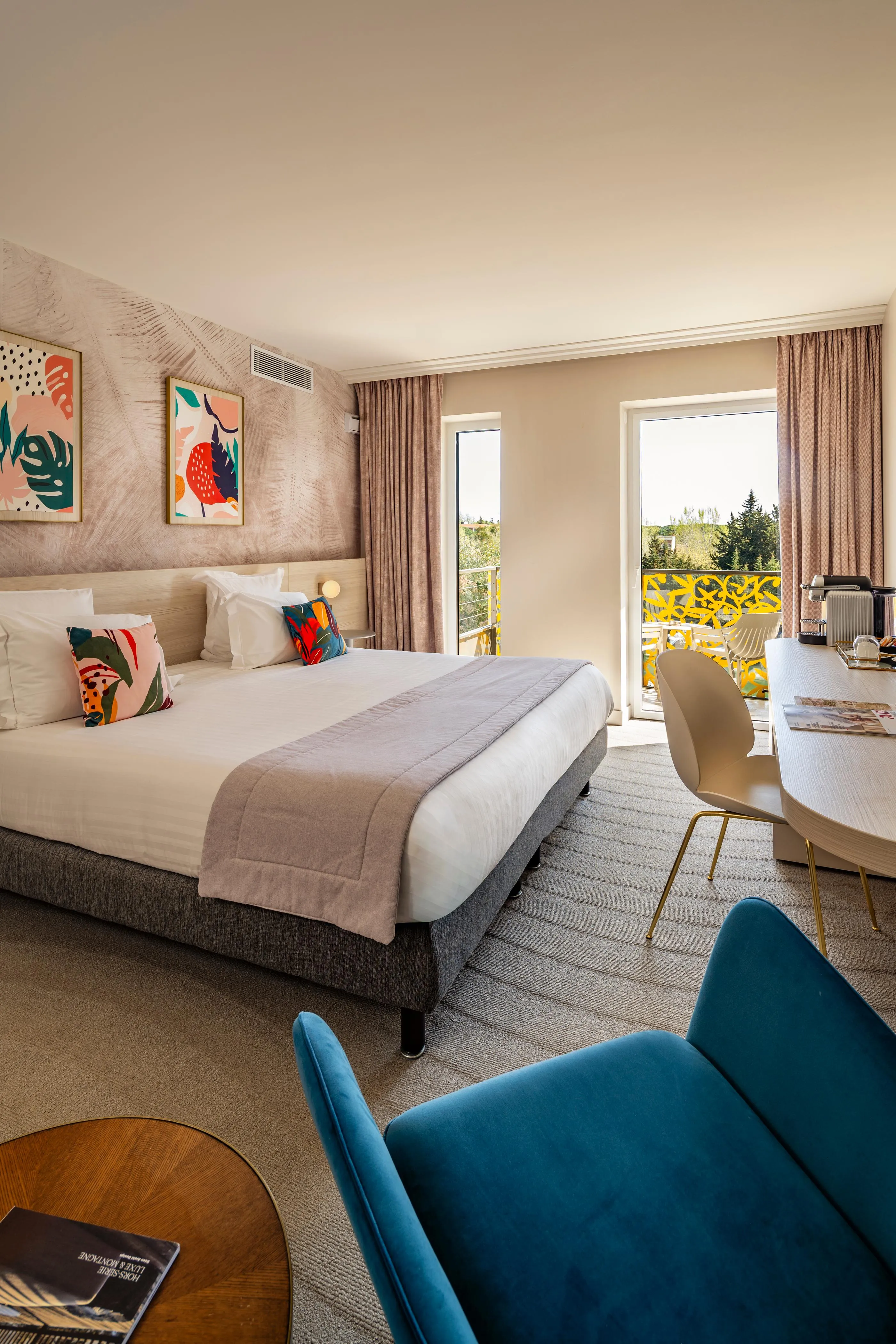 Hotel room with a bed, colorful cushions, two armchairs, a table with a chair, and a French window leading to a balcony.