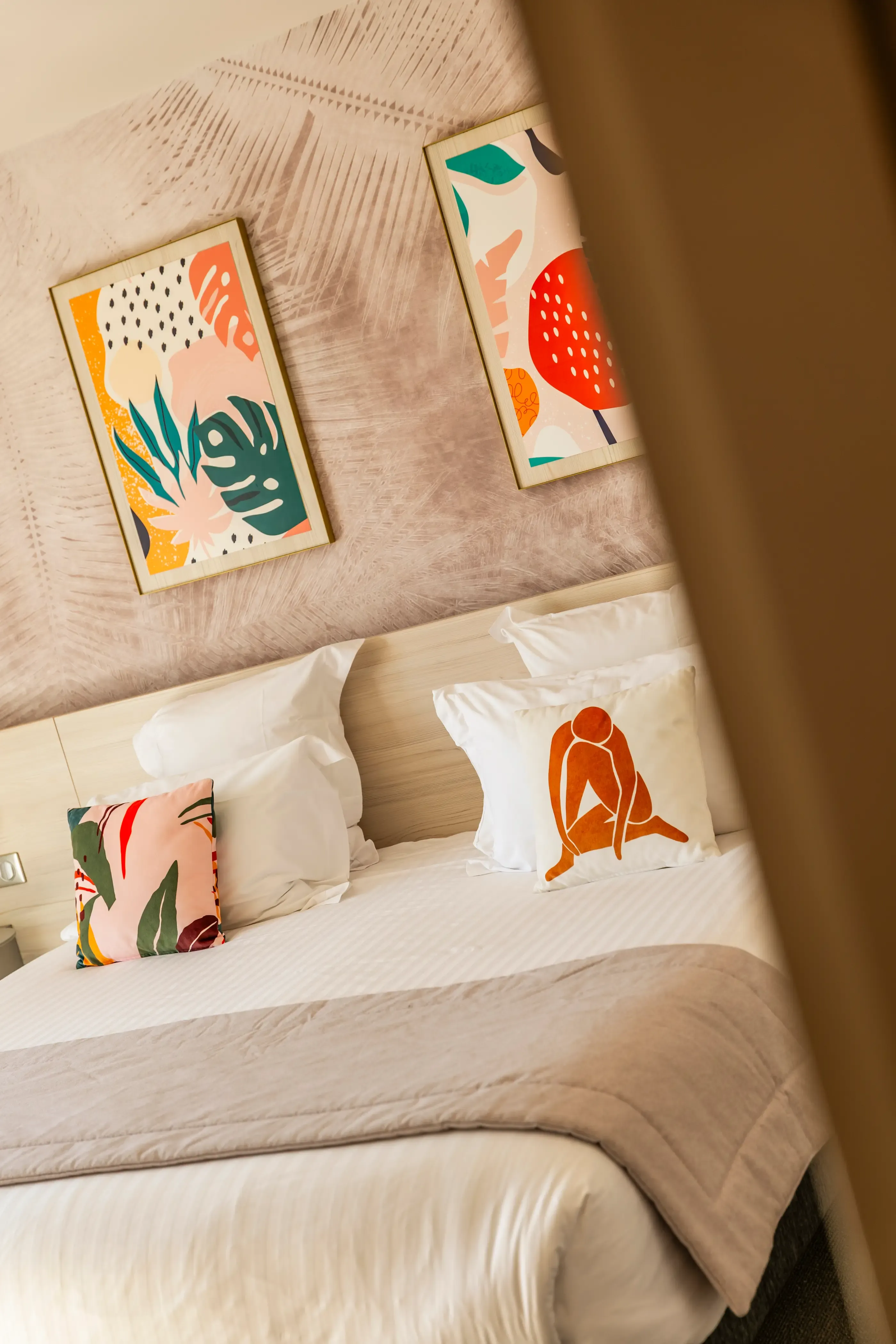 A bed with a light wood headboard, colorful cushions, and two abstract paintings hung on the wall.