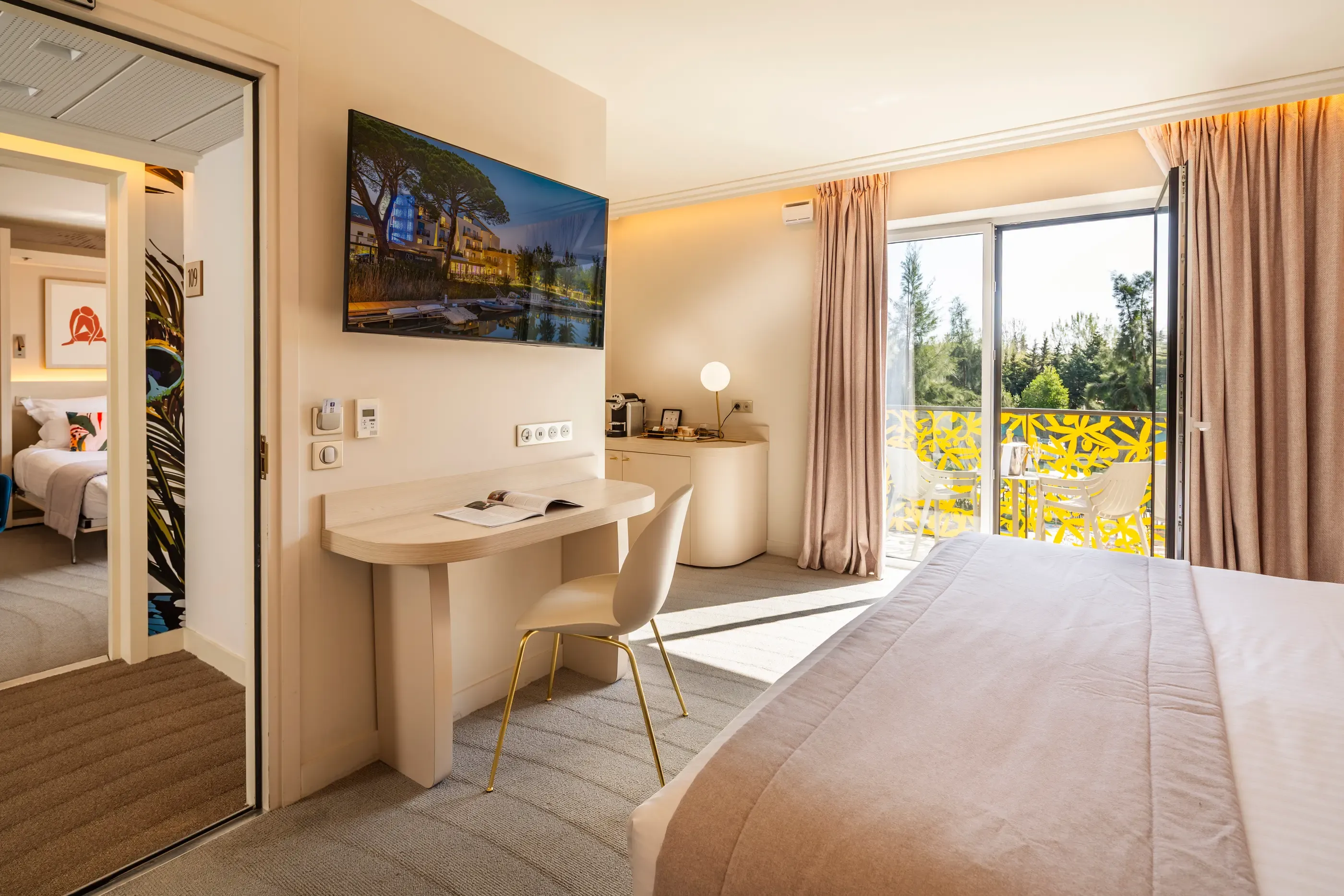 Hotel room with a bed, a desk, a television, and a French window opening onto a balcony with a view of trees.