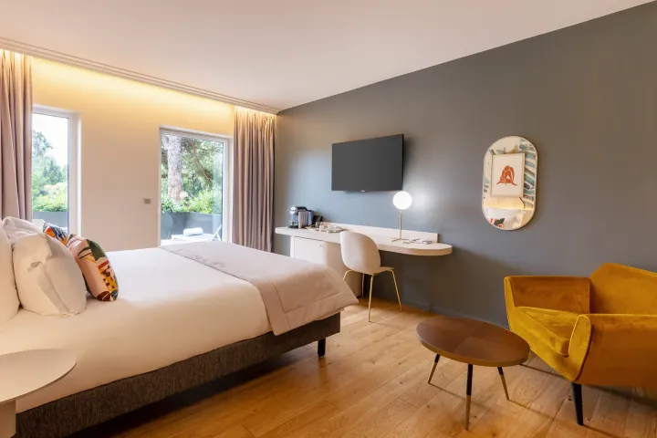 Chambre moderne avec un lit, une télévision, un bureau, un fauteuil jaune et une table basse, éclairée par la lumière naturelle des fenêtres