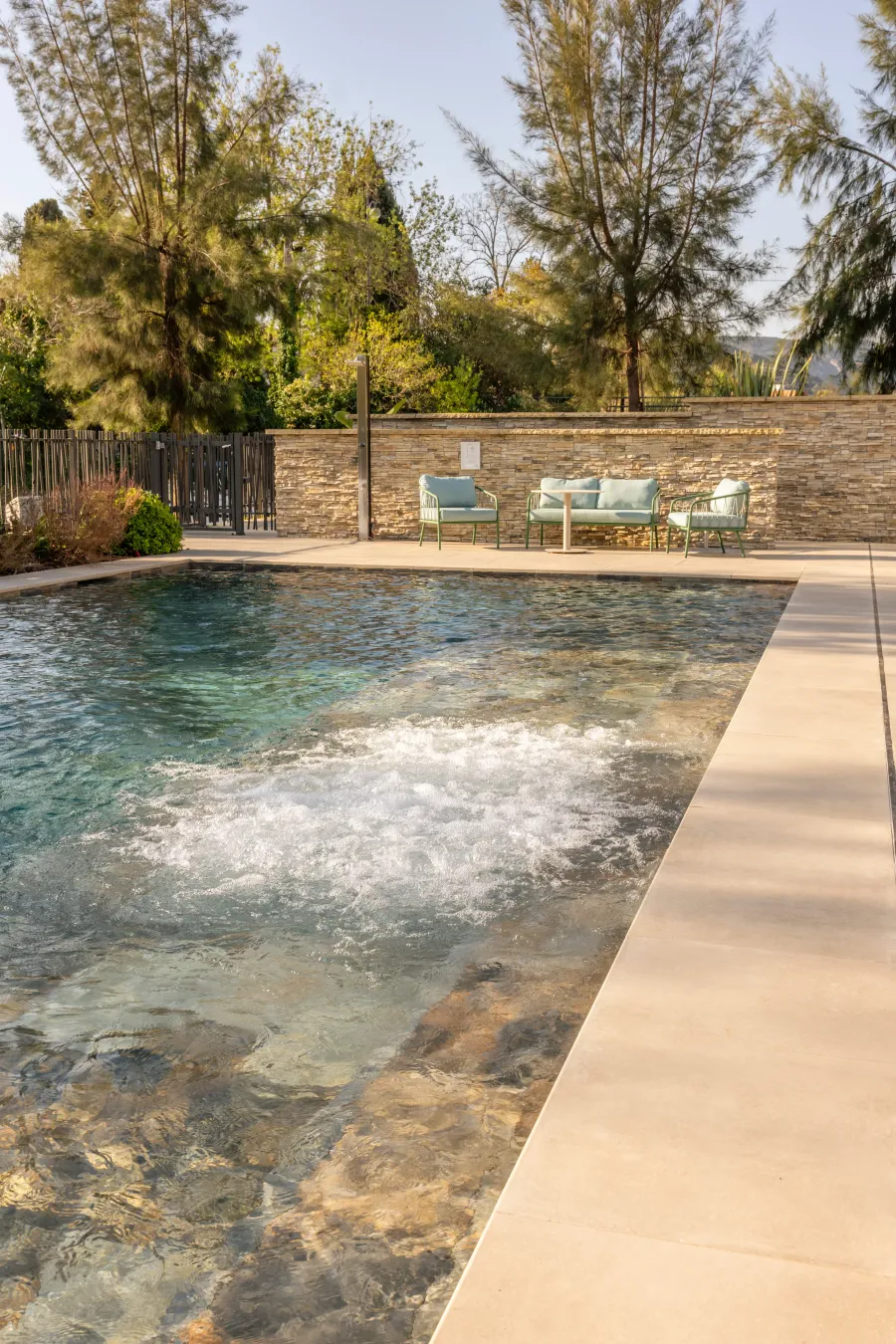 Pool with a relaxation area consisting of lounge chairs and a low table, surrounded by trees and a stone fence.