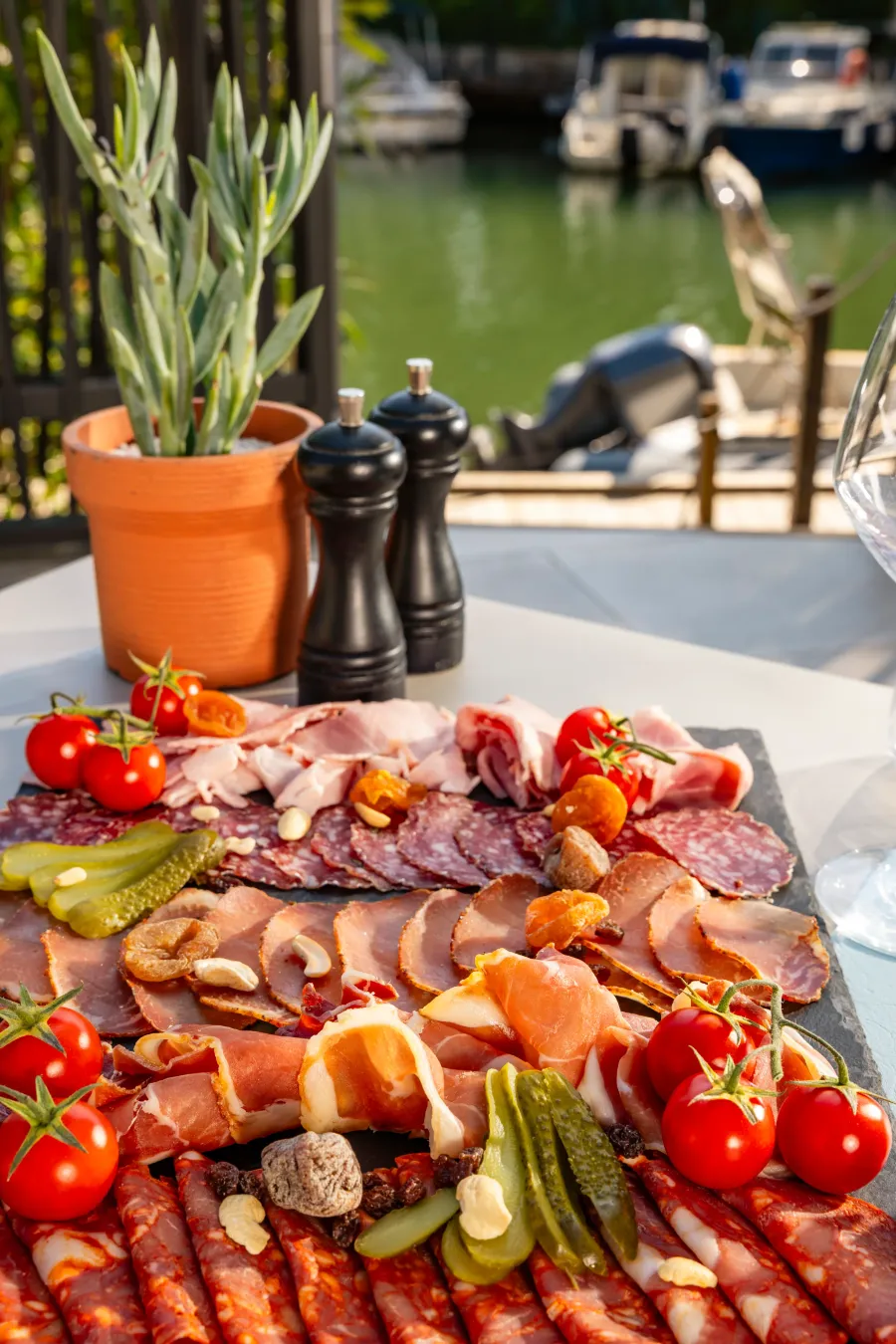 A charcuterie board with various slices of meat, cherry tomatoes, pickles, and nuts, placed on an outdoor table near a body of water.