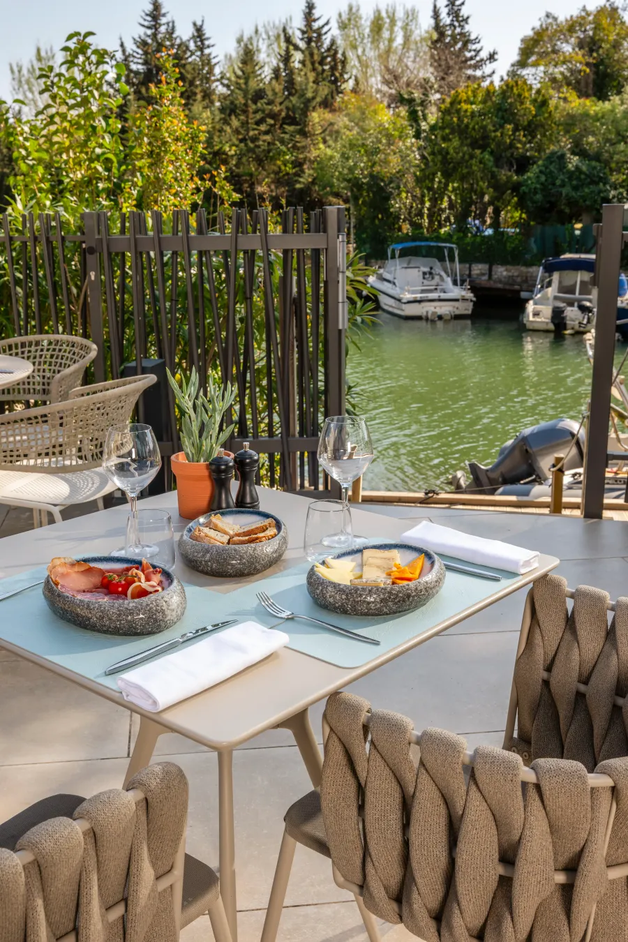Table set outdoors with dishes, glasses, and cutlery, facing a canal with moored boats and trees in the background.
